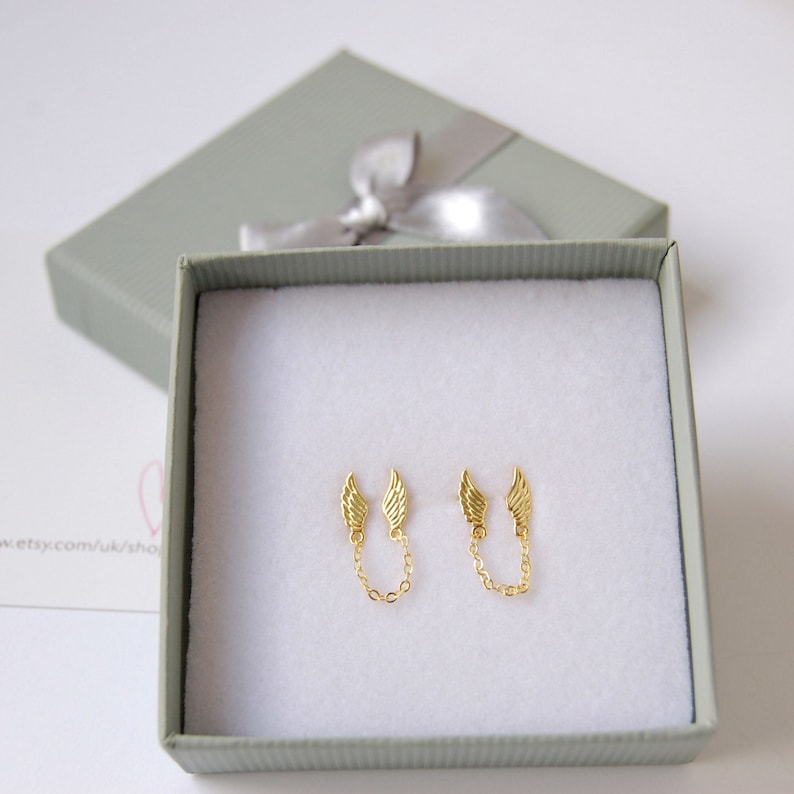 May include: A pair of gold-tone earrings with a delicate chain connecting two small wing charms. The earrings are displayed in a white box with a gray lid.