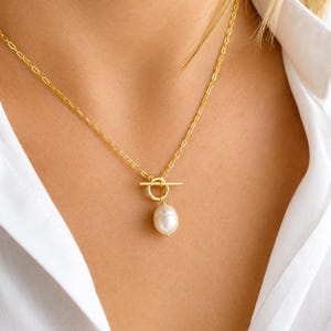 May include: A gold necklace featuring a toggle clasp and a single, white, pear-shaped pearl pendant. The necklace is set against a white shirt.