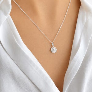 May include: A silver-toned necklace featuring a delicate chain and a small, multi-petaled flower-shaped pendant. The pendant is composed of several tiny, clear, reflective stones arranged in a circular pattern, creating a bright, sparkling effect.