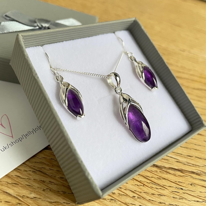 Op de afbeelding: Een zilveren ketting en oorbellen set met paarse amethist stenen. De stenen zijn gezet in een delicaat zilveren ontwerp. De sieraden worden getoond in een grijze doos.