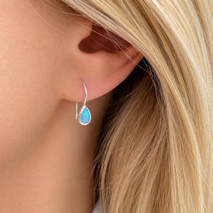 May include: A silver teardrop earring with a blue opal stone is shown. The earring is hanging from the ear. The opal stone has a pear shape and is surrounded by a silver setting. The earring is a simple and elegant design.