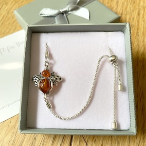 May include: A silver chain necklace with a bee charm featuring an amber stone. The necklace is in a white gift box with a grey lid.