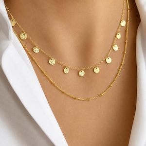 May include: A layered gold necklace featuring two distinct chains. The upper chain is adorned with small, round, textured gold discs spaced evenly along the length. The lower chain is a delicate gold cable chain with small, evenly spaced gold beads.