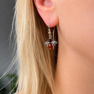 May include: A silver-toned dangle earring shaped like a bee, featuring a dark amber-colored stone for the body and intricate metal filigree wings. The earring hangs from a simple silver hook, showcasing a detailed insect design with warm tones.