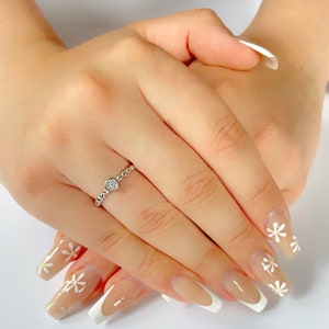 May include: A silver ring with a small round diamond-like stone set in the center. The ring is on a finger with a light brown nail polish and a white daisy design.