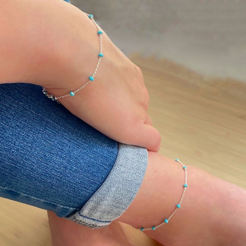 Op de afbeelding: Een zilveren enkelband- en armbandset met kleine, ronde turquoise kralen. De fijne sieraden zijn geregen op een fijne ketting. De enkelband wordt om de enkel gedragen en de armband om de pols. Lichte achtergrond.