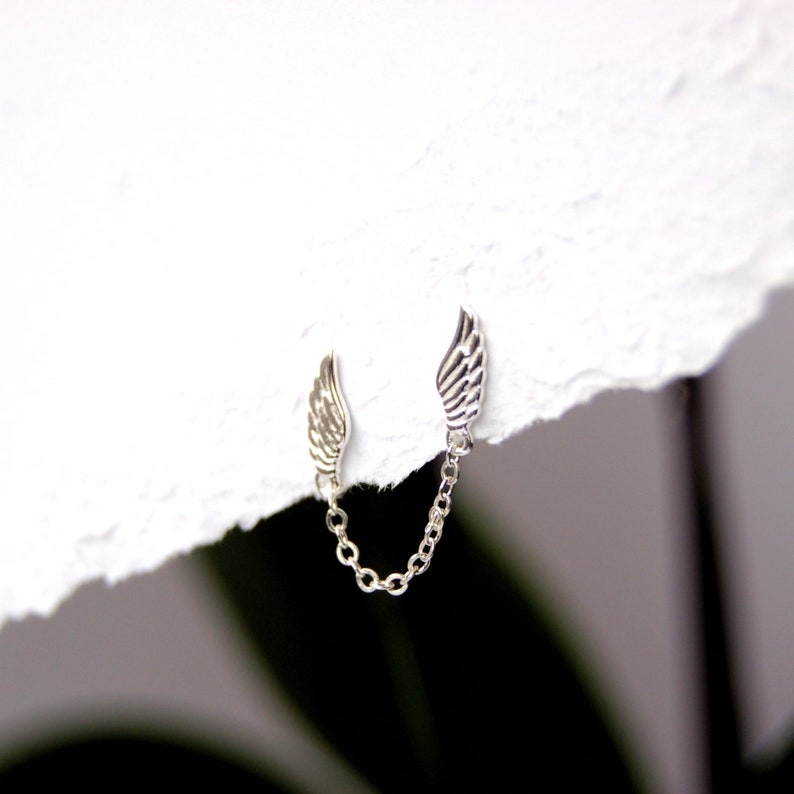 May include: A pair of silver wing earrings connected by a silver chain. The wings are detailed and have a delicate design.