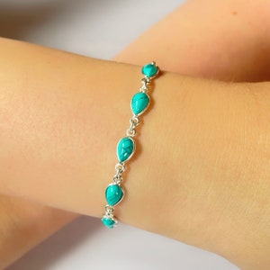 Op de afbeelding: Een zilveren armband met turkooizen edelstenen. De armband heeft een reeks verbonden, druppelvormige turkooizen stenen, afgewisseld met kleine ronde turkooizen stenen. De armband wordt om een pols gedragen.