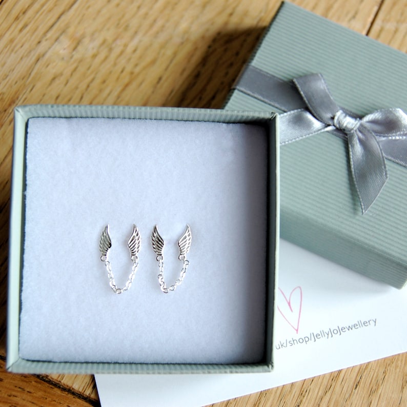 May include: A pair of silver earrings with delicate angel wings connected by a chain. The earrings are displayed in a small white box with a gray lid. The box is sitting on a wooden surface.