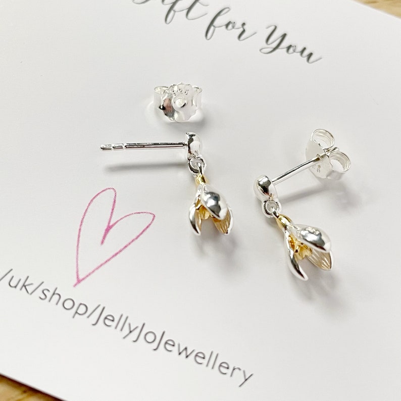 Op de afbeelding: Een paar zilveren oorbellen met een gouden bloementje. De oorbellen liggen op een witte kaart met de tekst "Gift for you" en "uk/shop/jellyjojewellery".