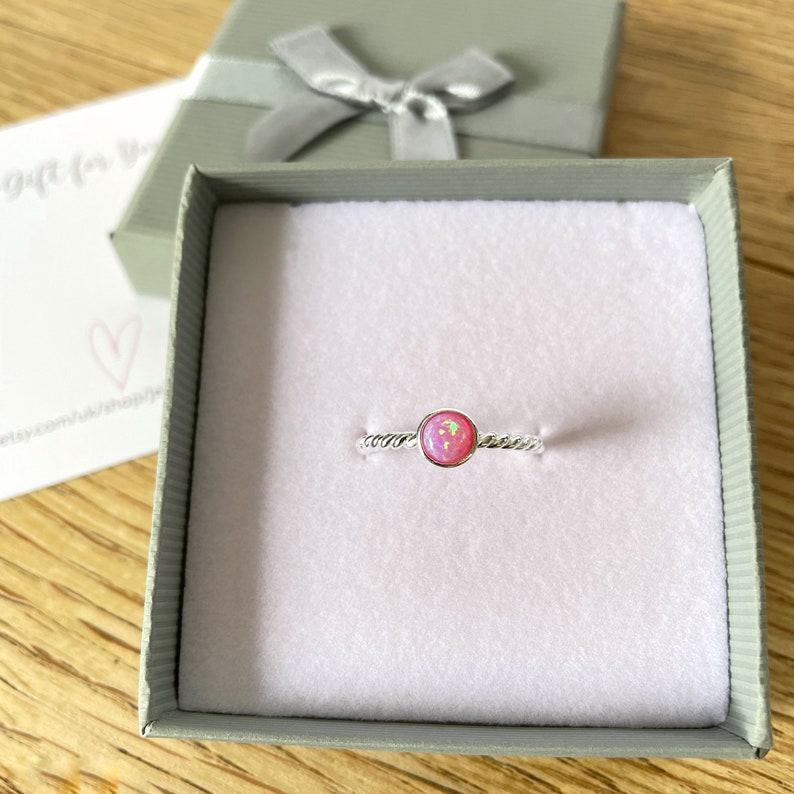 May include: A silver ring with a pink opal gemstone set in a twisted band. The ring is displayed in a white gift box.