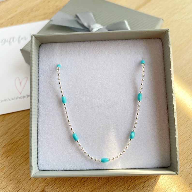 May include: A delicate silver necklace with turquoise beads, presented in a gray gift box with a silver bow. The necklace features small silver beads interspersed with oval turquoise beads. A card with a heart and text is visible.