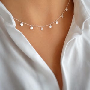 Puede incluir: Un delicado collar de cadena plateada que presenta múltiples dijes circulares pequeños, planos y espaciados uniformemente a lo largo de la cadena. El collar descansa sobre el cuello, usado con una camisa blanca de cuello abierto.