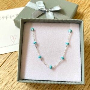 Op de afbeelding: Een delicate zilveren ketting met turquoise kralen wordt gepresenteerd in een grijze geschenkdoos met een zilveren lint. De ketting ligt op een lichtroze ondergrond, met een kleine kaart op de achtergrond.