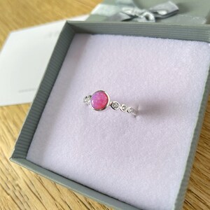 Op de afbeelding: Een zilveren ring met een roze opaal-edelsteen en kleinere heldere edelstenen die in een band zijn gezet.