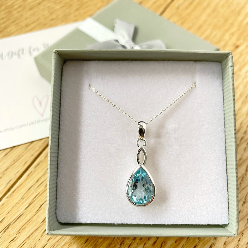 May include: A silver necklace with a teardrop-shaped blue gemstone pendant. The pendant is set in a silver setting and hangs from a delicate silver chain. The necklace is displayed in a small, white gift box.