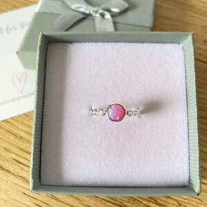 Op de afbeelding: Een zilveren ring met een roze opaal-steen en kleine heldere stenen rond de band. De ring zit in een witte doos.