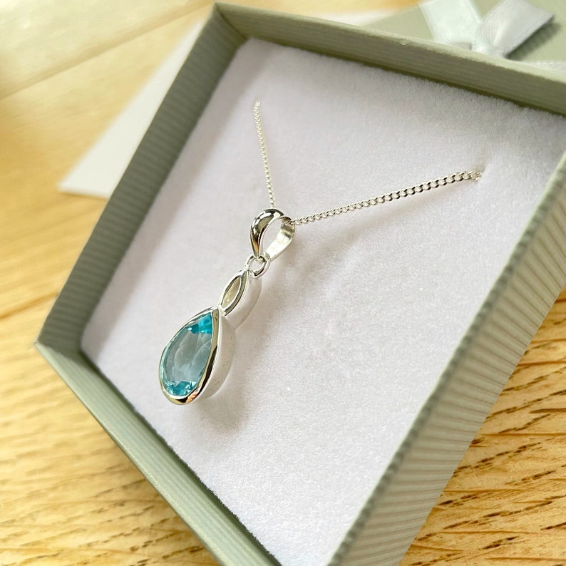 May include: A silver necklace with a teardrop-shaped blue gemstone pendant. The necklace is in a white box with a grey lid.