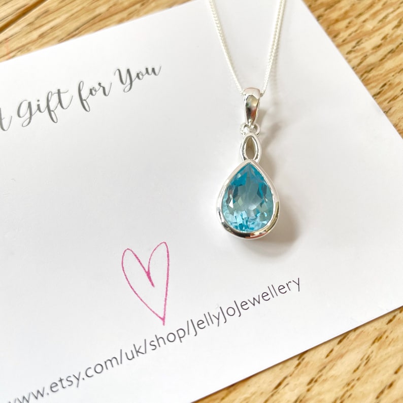 May include: A silver necklace with a teardrop-shaped pendant featuring a light blue gemstone. The pendant has a delicate silver design around the gemstone. The necklace is displayed on a white card with the text "Gift for You" and a website address.