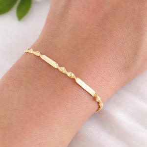 Puede incluir: Una delicada pulsera dorada con un diseño texturizado y ondulado. La pulsera presenta una combinación de eslabones planos y rectangulares y segmentos retorcidos en forma de cuerda. La joya se lleva en la muñeca.