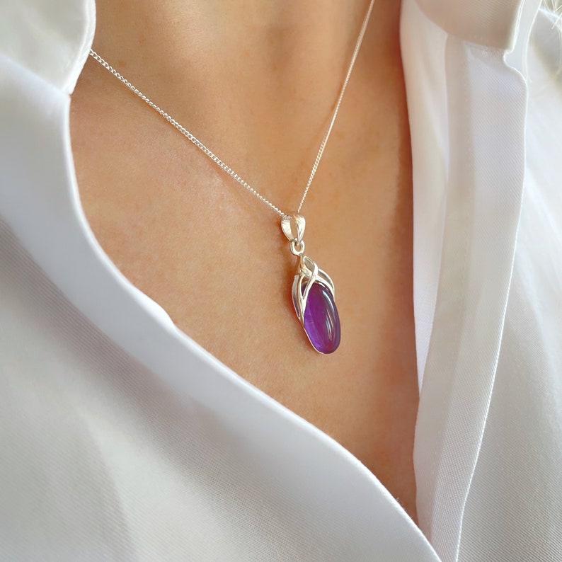 Op de afbeelding: Een zilveren ketting met een hanger van een paarse ovale edelsteen. De hanger is gezet in een zilveren frame met een delicaat ontwerp.