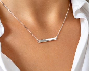 May include: A polished silver-tone bar pendant necklace featuring a slender, horizontal rectangular metal bar suspended from a delicate, fine-link chain. The smooth, reflective surface of the bar catches the light, creating a clean and simple aesthetic.