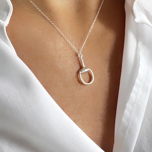 May include: A silver-colored necklace featuring a horse bit pendant. The pendant has a ring and a bar design. The necklace is displayed against a white shirt.