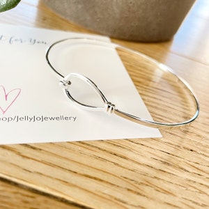 May include: A silver bangle bracelet with a unique teardrop-shaped design. The bracelet is open and has a simple, elegant look.