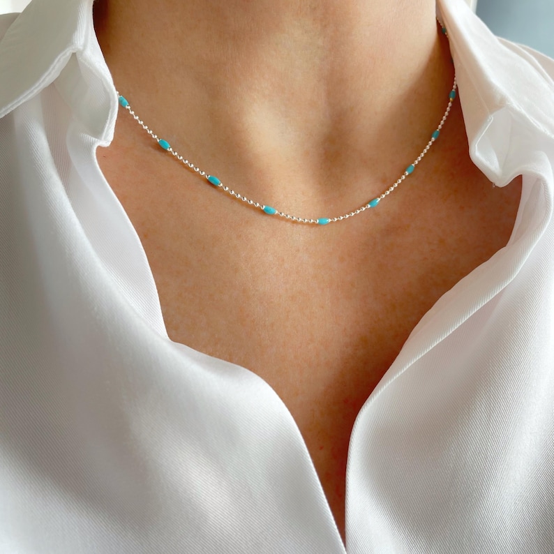 May include: A delicate silver necklace featuring small, turquoise-colored beads evenly spaced along a fine chain. The necklace is shown against a white collared shirt, highlighting the jewelry's subtle elegance. This necklace is a perfect accessory for everyday wear.