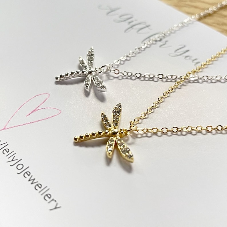 May include: Two dragonfly necklaces, one in silver and one in gold, each featuring clear stones on the wings and a beaded tail. The necklaces are displayed on a white card with the text A gift for you and jellyjojewellery with a pink heart.