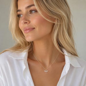 May include: A silver-toned necklace featuring a delicate dragonfly pendant with small, clear, sparkling stones embedded along the wings and body, suspended from a thin, simple metal chain worn around the neck against a plain white shirt.