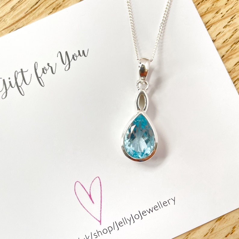 May include: A silver necklace featuring a teardrop-shaped blue topaz pendant. The pendant is set in a silver bezel and hangs from a delicate silver chain. The necklace is displayed on a white card with the text "Gift for You" and a pink heart.