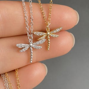 May include: Two dragonfly necklaces, one in silver tone and one in gold tone, each featuring a delicate chain and a pendant encrusted with small clear stones along the wings. The dragonfly bodies are composed of a series of small metal spheres.