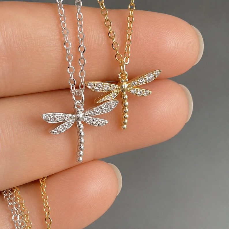 May include: Two dragonfly necklaces, one in silver tone and one in gold tone, each featuring a delicate chain and a pendant encrusted with small clear stones along the wings. The dragonfly bodies are composed of a series of small metal spheres.