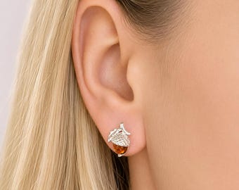 Puede incluir: Un pendiente de plata con forma de bellota, que presenta una tapa de plata texturizada y una base de piedra translúcida de color ámbar. El pendiente está sujeto al lóbulo de la oreja, mostrando un acabado metálico y una piedra cálida.