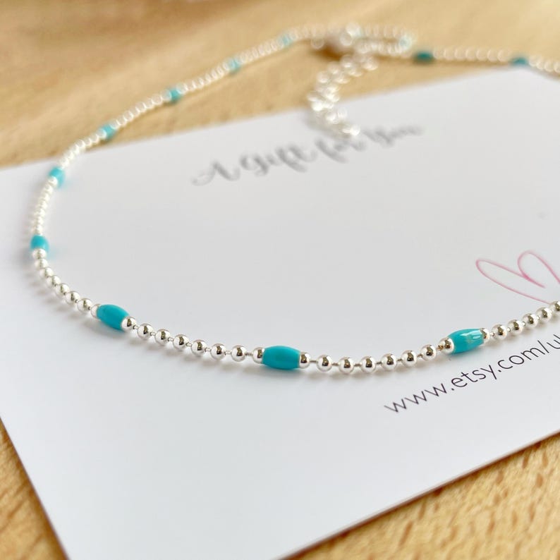 May include: A delicate silver necklace with small, round beads and vibrant turquoise-colored oval beads. The necklace is displayed on a white card with the website address www.etsy.com/u...