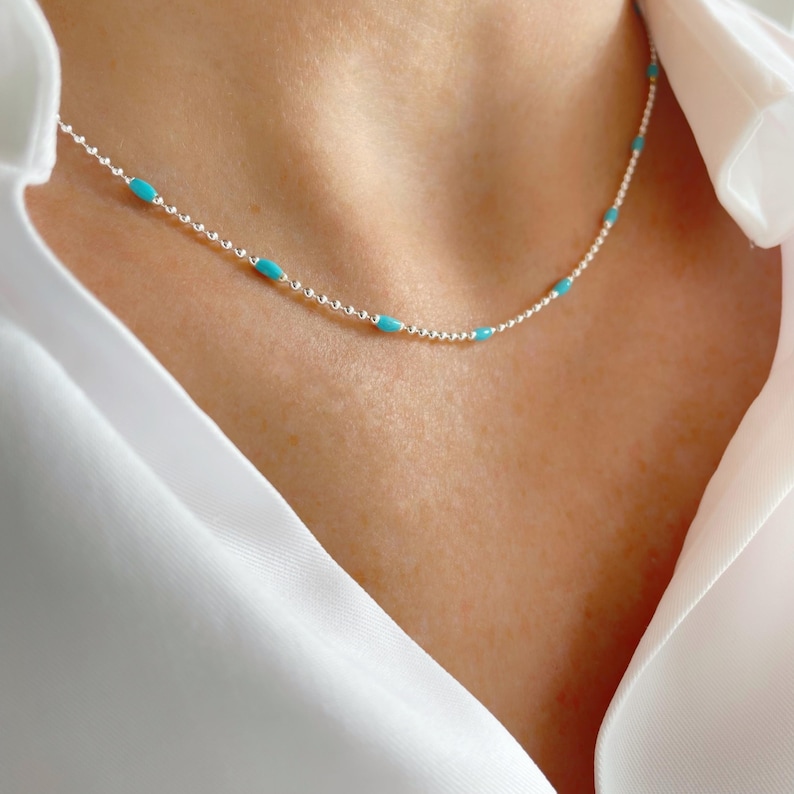 May include: A delicate silver necklace with small, turquoise-colored beads evenly spaced along the chain. The necklace is worn against a person's skin, with a white shirt in the background.