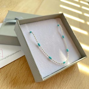 May include: A delicate silver necklace with turquoise-colored beads, presented in a gray gift box with a ribbon. The necklace is displayed on a white surface inside the box, with a shop card visible.