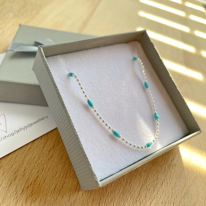 May include: A delicate silver necklace with turquoise-colored beads, presented in a gray gift box with a ribbon. The necklace is displayed on a white surface inside the box, with a shop card visible.