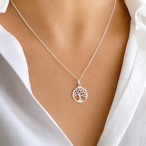 Könnte beinhalten: Eine silberfarbene Halskette mit einem runden Anhänger, der ein ausgeschnittenes Baummotiv zeigt. Der Anhänger hängt an einer zarten, dünnen silberfarbenen Kette, die auf dem Schlüsselbein aufliegt. Das Metall hat eine polierte Oberfläche.