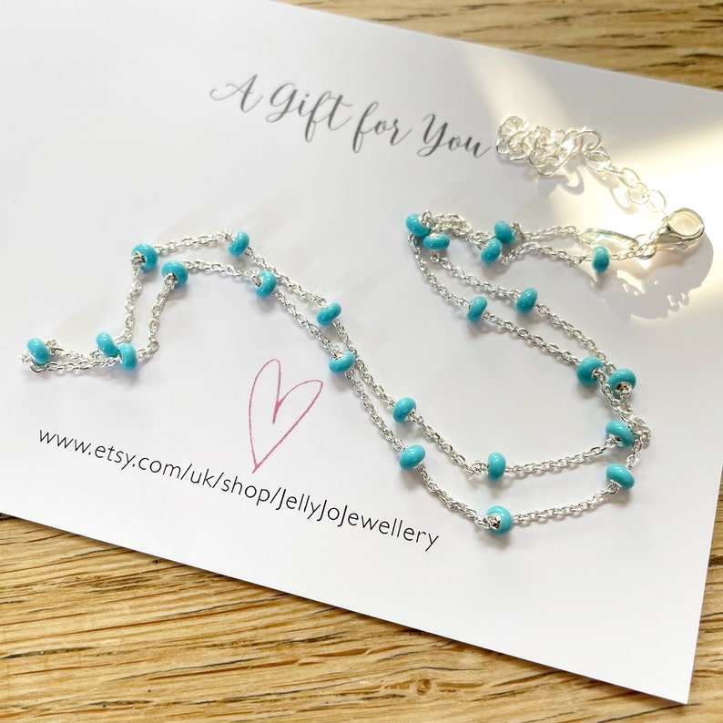 Op de afbeelding: Een delicate zilveren ketting met kleine, turquoise kralen. De ketting ligt op een witte kaart met de tekst "A Gift for You" en een hart. De Etsy shop URL is zichtbaar.