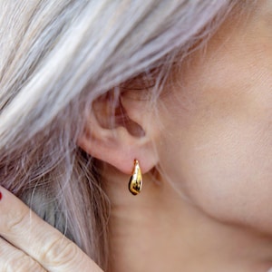 May include: A pair of gold hoop earrings with a teardrop shape. The earrings are worn in the ear of a person with blonde hair.