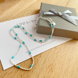 May include: A silver necklace and bracelet set with turquoise-colored beads. The jewelry is displayed on a card that reads "A Gift for You" and a gray gift box with a silver ribbon.