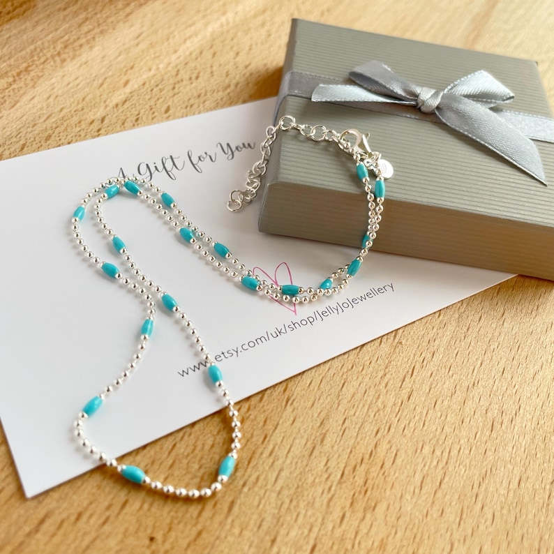 May include: A silver necklace and bracelet set with turquoise-colored beads. The jewelry is displayed on a card that reads "A Gift for You" and a gray gift box with a silver ribbon.
