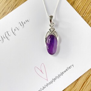 Op de afbeelding: Een zilveren ketting met een hanger van paarse amethist. De hanger is gezet in een zilveren swirl-ontwerp. De ketting ligt op een witte kaart met de tekst "Cadeau voor jou" en een roze hart.