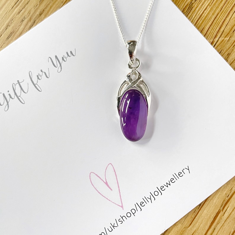 Op de afbeelding: Een zilveren ketting met een hanger van paarse amethist. De hanger is gezet in een zilveren swirl-ontwerp. De ketting ligt op een witte kaart met de tekst "Cadeau voor jou" en een roze hart.