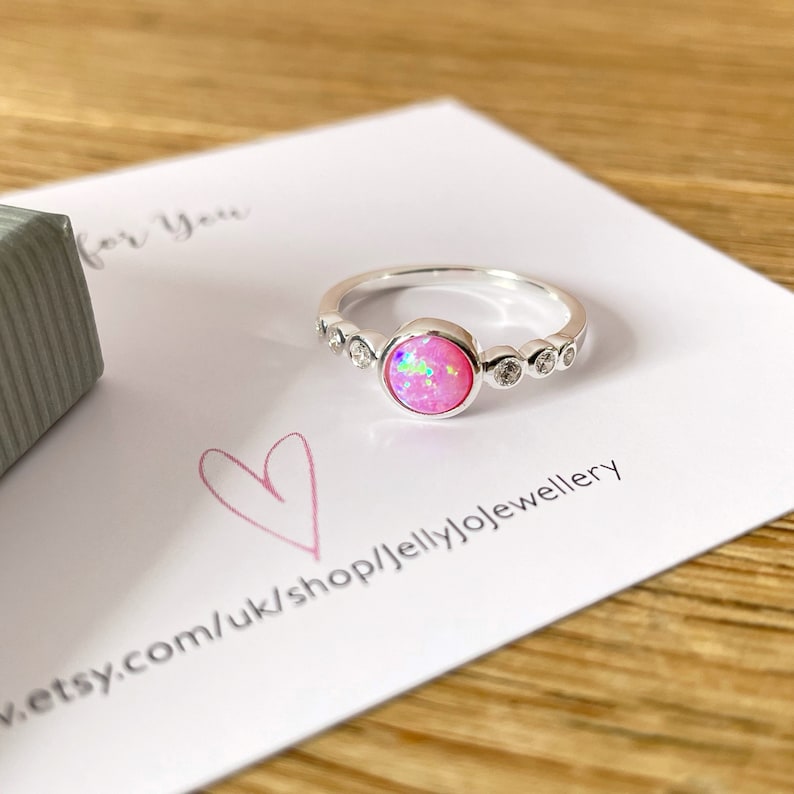 Op de afbeelding: Een zilveren ring met een roze opaal edelsteen en kleine heldere stenen rond de band. De ring ligt op een witte kaart met de tekst 'for you' en een websiteadres.