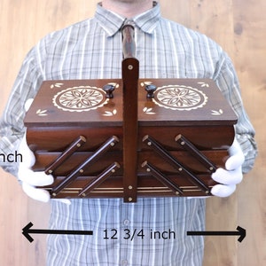 May include: A dark brown wooden accordion-style sewing box with a central handle, two lids featuring white floral inlays, and a tiered design. The box measures 6 1/4 inches in height and 12 3/4 inches in width, with the text LINDENBOXES visible.