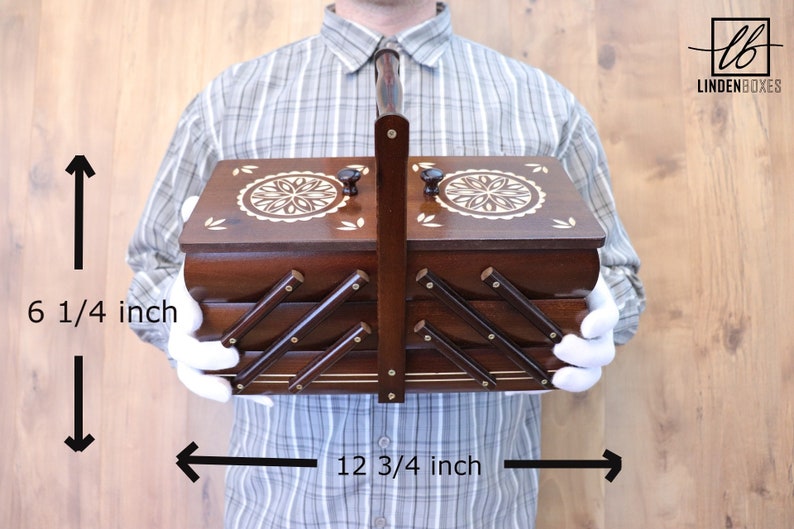 May include: A dark brown wooden accordion-style sewing box with a central handle, two lids featuring white floral inlays, and a tiered design. The box measures 6 1/4 inches in height and 12 3/4 inches in width, with the text LINDENBOXES visible.