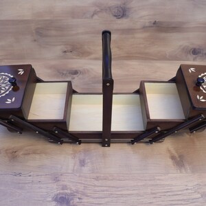 May include: A dark brown wooden sewing box with a cantilevered design, featuring two hinged lids with light-colored floral inlay patterns, a central vertical handle, and multiple open storage compartments with light-colored interior surfaces.